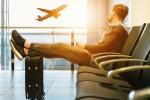 mężczyzna siedzący na fotelu na lotnisku, trzyma w ręku komórkę, nogi ma na walizce, spogląda za okno na lecący samolot